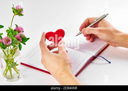 caucasian person taking notes at dairy and holding red heart at his arm over white background. summer romance notes concept Stock Photo