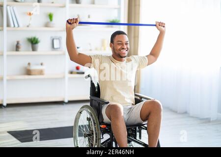 Happy black disabled man in wheelchair doing exercises with rubber band at home Stock Photo