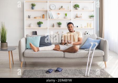 Millennial black guy with plastered leg sitting on sofa, reading book at home, pair of crutches standing nearby Stock Photo