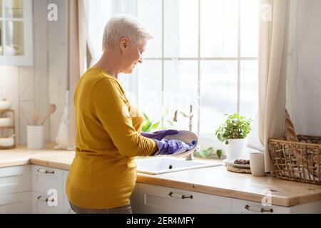 Senior lady washing dishes in kitchen, using rubber gloves Stock Photo