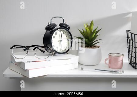 A stack of books, an artificial succulent plant in a ceramic pot, glasses, a metal basket with notebooks, a pen and a cup on a white table surface. Co Stock Photo