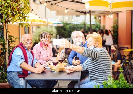 Senior couples drink red wine at bar with face masks protection - New normal lifestyle concept with happy people having fun together taking a selfie Stock Photo