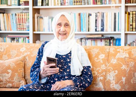 Happy old arabic muslim woamn using technology while sitting on couch Stock Photo