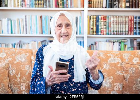 Happy old arabic muslim woamn using technology while sitting on couch Stock Photo