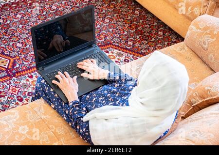 Happy old arabic muslim woamn using technology while sitting on couch Stock Photo