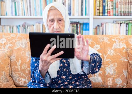 Happy old arabic muslim woamn using technology while sitting on couch Stock Photo