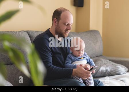 Caucasian father talking on mobile phone to work online with his newborn baby. Single dad taking care of his son. Happy family and work at home Stock Photo