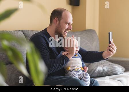 Happy father and son taking selfie with smartphone sitting on the couch at home. Portrait smiling Dad and child. Child making video call with mobile Stock Photo