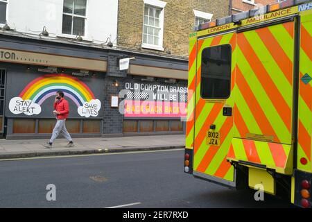 A man wearing a facemask as a precaution against the spread of covid 19, walks opposite a 'Stay Alert, Say Lives' graffiti, and an emergency ambulance parked  across the street in London. Stock Photo