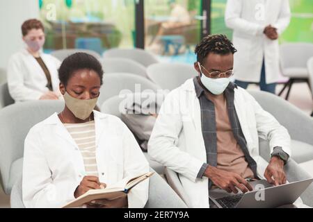 Portrait of young African American woman wearing mask and lab coat while listening to lecture on medicine in college or coworking center, copy space Stock Photo