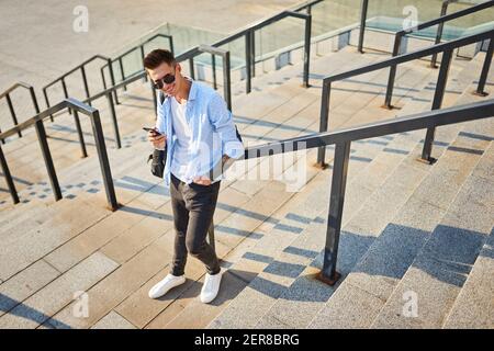 Young man in sunglasses and with a black backpack. Standing on the steps with the phone in hand and smiling looks into the frame Stock Photo