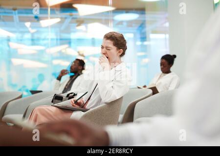 Portrait of young woman yawning while sitting in audience and listening to lecture on medicine in college or coworking center, copy space Stock Photo