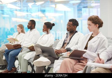 Multi-ethnic group of people wearing lab coats sitting in row in audience at medical seminar, copy space Stock Photo