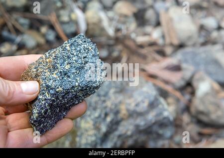 Fingers holding piece of chromite ore over natural rocks background, soft focus Stock Photo