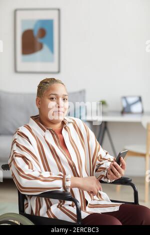 Vertical portrait of modern mixed-race woman sitting in wheelchair and using smartphone while looking at camera in minimal home interior, copy space Stock Photo
