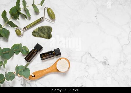 Natural skin care, spa and wellness tools: beauty jade roller, gua sha stone, essential oil and wooden face brush with eucalyptus branch on marble bac Stock Photo
