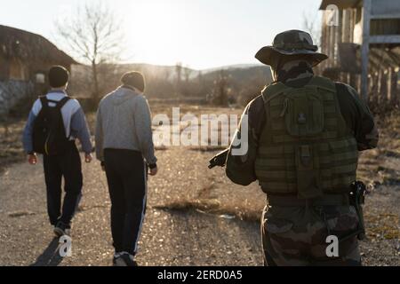 Group of two migrants held by soldier at border patrol on the road in nature in sunny day - Back view of two unknown men facing armed man in uniform o Stock Photo