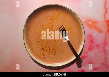 Dirty pink ceramic plate with food leftover on a pink grunge background. Empty shallow plate with fork. Background of the table after the end of lunch Stock Photo