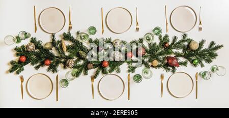 Flat-lay of festive Christmas table setting with dinnerware, cutlery, glassware, fur tree branches, candles, baubles and toys over white table backgro Stock Photo