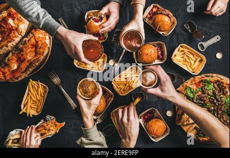 Family lockdown fast-food dinner from delivery service at home. Flat-lay of table with burgers, fries, sandwiches, beer and pizza and hands of people Stock Photo