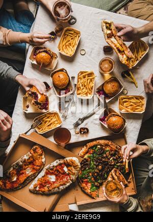 Fast food dinner from delivery service. Flat-lay of friends or relatives hands having quarantine home party with burgers, fries, sandwiches, pizza, be Stock Photo