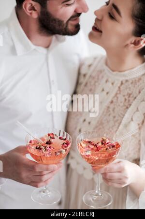 Happy romantic young caucasian couple looking at each other, hugging, holding glasses of cold sparkling champagne cocktail with rose petals and drinki Stock Photo