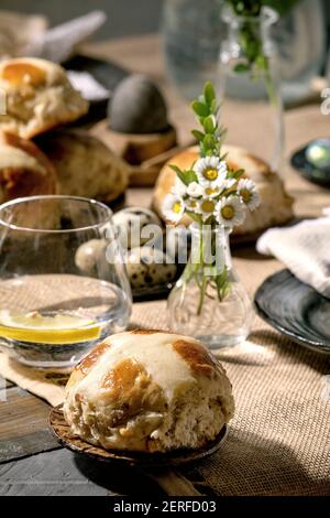 Easter table setting with colored and chocolate eggs, hot cross buns, bouquet flowers, empty ceramic plate with napkin, glass of lemonade drink on woo Stock Photo