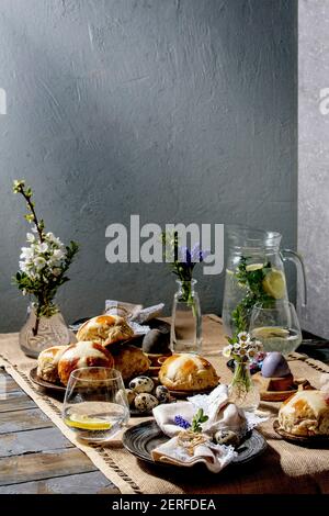 Easter table setting with colored and chocolate eggs, hot cross buns, bouquet flowers, empty ceramic plate with napkin, glass of lemonade drink on woo Stock Photo