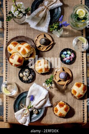Easter table setting with colored and chocolate eggs, hot cross buns, bouquet flowers, empty ceramic plate with napkin, glass of lemonade drink on woo Stock Photo
