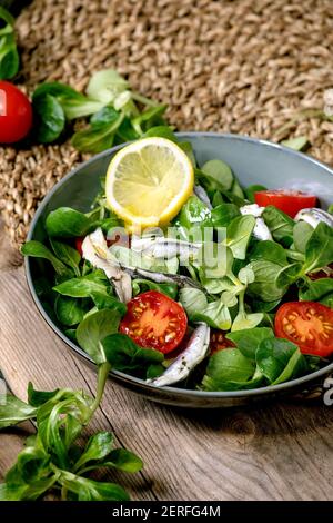 Green field salad with pickled anchovies or sardines fillet, and cherry tomatoes, served in blue bowl with lemon and olive oil on straw napkin over ol Stock Photo