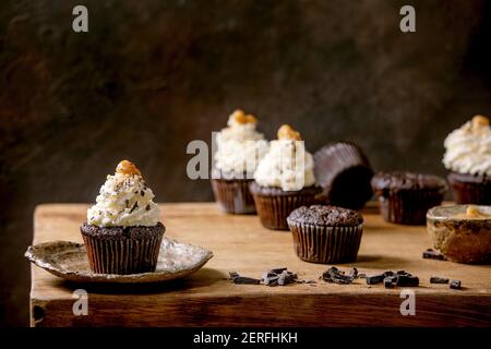 Homemade chocolate cupcakes muffins with white whipped butter cream and salted caramel on ceramic plate, served with chopped dark chocolate on wooden Stock Photo