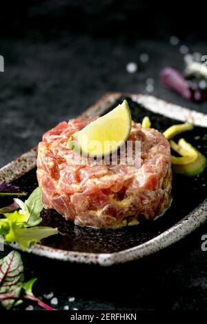 Tuna tartare with green salad, lime, avocado and mustard sauce serving on japanese style black ceramic plate over black marble background. Restaurant Stock Photo