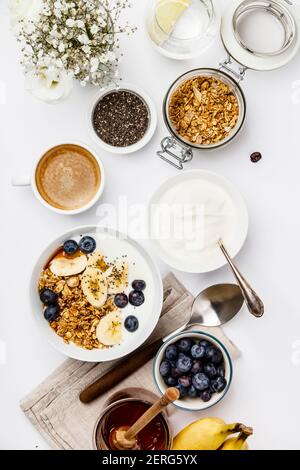 Healthy breakfast set. Oat granola with yogurt, honey, fresh bananas, blueberries, chia seeds in bowl  and cup of coffee on white background, top view Stock Photo