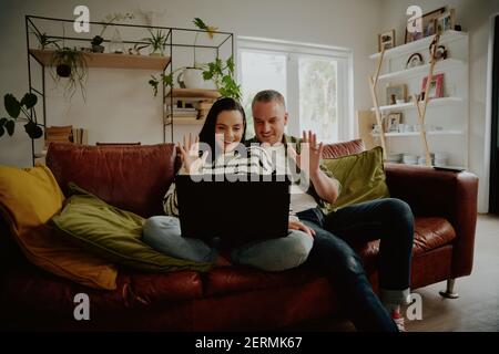Cheerful couple sitting on sofa and waving hands to laptop while chatting using video call at home Stock Photo