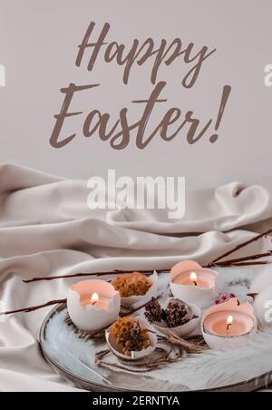 HAPPY EASTER lettering greeting card text. Easter DIY. Do it yourself. Trendy Easter decorations. Cracked organic eggs with dry grass and dry flowers on plate. Candle in White shell of the eggs. Bird feather. Pastel colors. Stock Photo