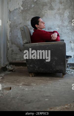 Sad woman with short hair sitting on armchair inside a derelict building Stock Photo