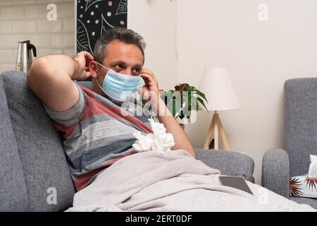 Ill adult man with sars covid symptoms wearing surgical or medical disposable mask to prevent contamination at home covering nose and mouth sitting on Stock Photo
