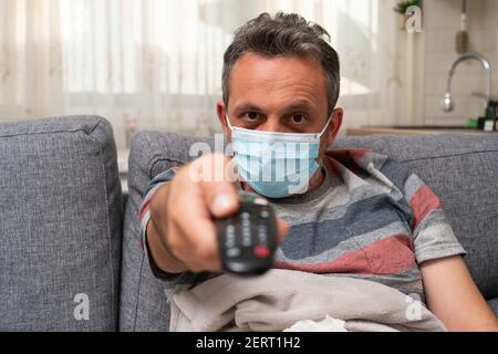 Close-up of sick man at home on sofa wearing protective disposable medical or surgical face mask zapping channels on tv using remote control as home p Stock Photo