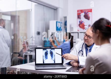 Doctor and nurse analising digital lung x-ray of patient sitting in medical office. Senior physician showing radiography result to young woman pointing with pancil on desktop discussing in front of pc