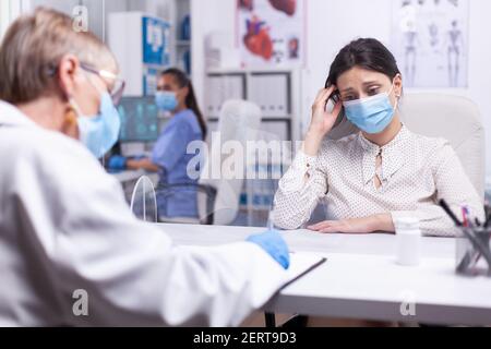 Young patient feeling sad receving bad news from senior doctor during covid-19 pandemic. The doctor telling woman about injury symptom and looking information in clipboard. Stock Photo