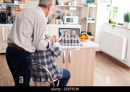 Senior couple during video conference with daughter in kitchen using laptop. Enthusiastic grandparents talking with family online using webcam during virtual discussion, modern communication online Stock Photo