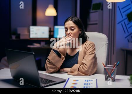 Exhausted businesswoman yawning working on important project in financial firm. Smart woman sitting at her workplace in the course of late night hours doing her job. Stock Photo