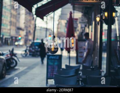 A Lightbulb hangs above a table of an urban café. The close up of the bulb on the street shows the bulb lit and in the background the waiter enjoys a Stock Photo