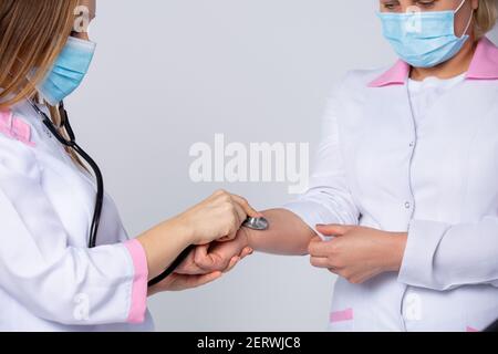 A young doctor in a white lab coat, wearing protective medical mask, is checking the pulse of her colleague- senior doctor with stethoscope. Empathy Stock Photo