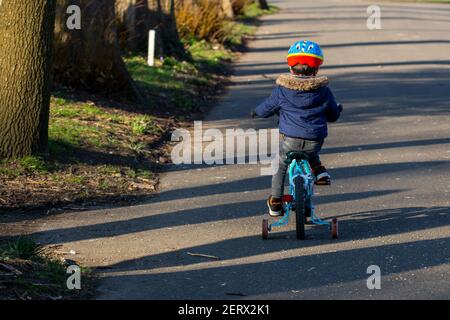 A small boy wearing a cycle helmet riding a bike in the park with stabilisers or stabilizers Stock Photo