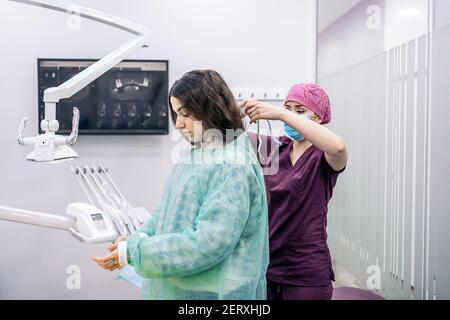 Stock photo of female dentist helping young patient to put on disposable clothes for surgery procedure. Stock Photo