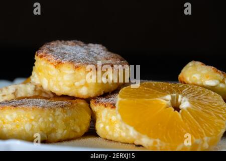 Sweet Cottage cheese pancakes with orange slices on a white plate close-up against a black wall with copy space. Stock Photo