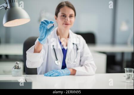 A young female doctor in blue medical gloves shows a patient a capsule with a new drug. Portrait on the background of the medical office Stock Photo