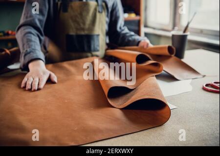 A young apprentice in a boot workshop prepares leather for further use on a large table Stock Photo