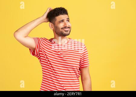 Confused uncertain man with beard in red striped t-shirt scratching his head, choosing, trying to make right decision, dilemma. Indoor studio shot iso Stock Photo